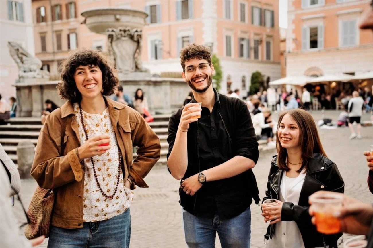 Three people smiling and holding drinks in a lively outdoor plaza with a fountain and historic buildings in the background.