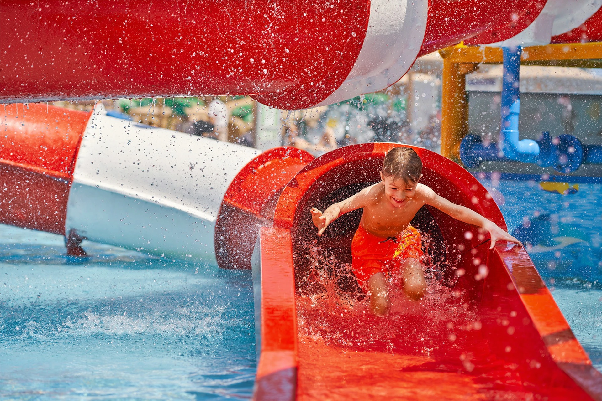 Child in red shorts exiting a red water slide, splashing into the pool below, with colorful slides and people in the background.