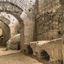 Ancient stone and brick archways and walls of a dimly lit, underground ruin or historic site with rough surfaces and vaulted ceilings.