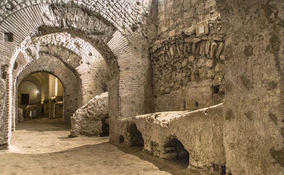 Ancient stone and brick archways and walls of a dimly lit, underground ruin or historic site with rough surfaces and vaulted ceilings.