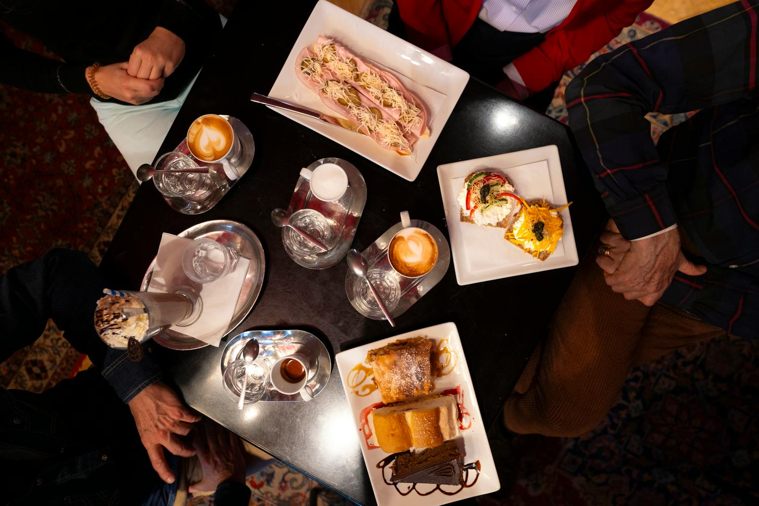 Uma mesa com vários pratos, incluindo sobremesas, sandes e café, rodeada por pessoas sentadas à mesa.