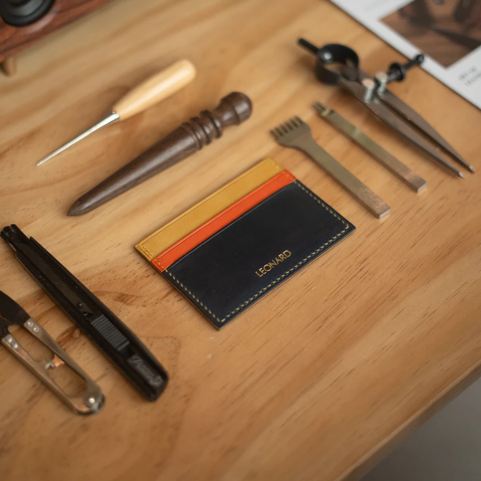A leather cardholder with orange, yellow, and navy sections on a wooden surface surrounded by various crafting tools.
