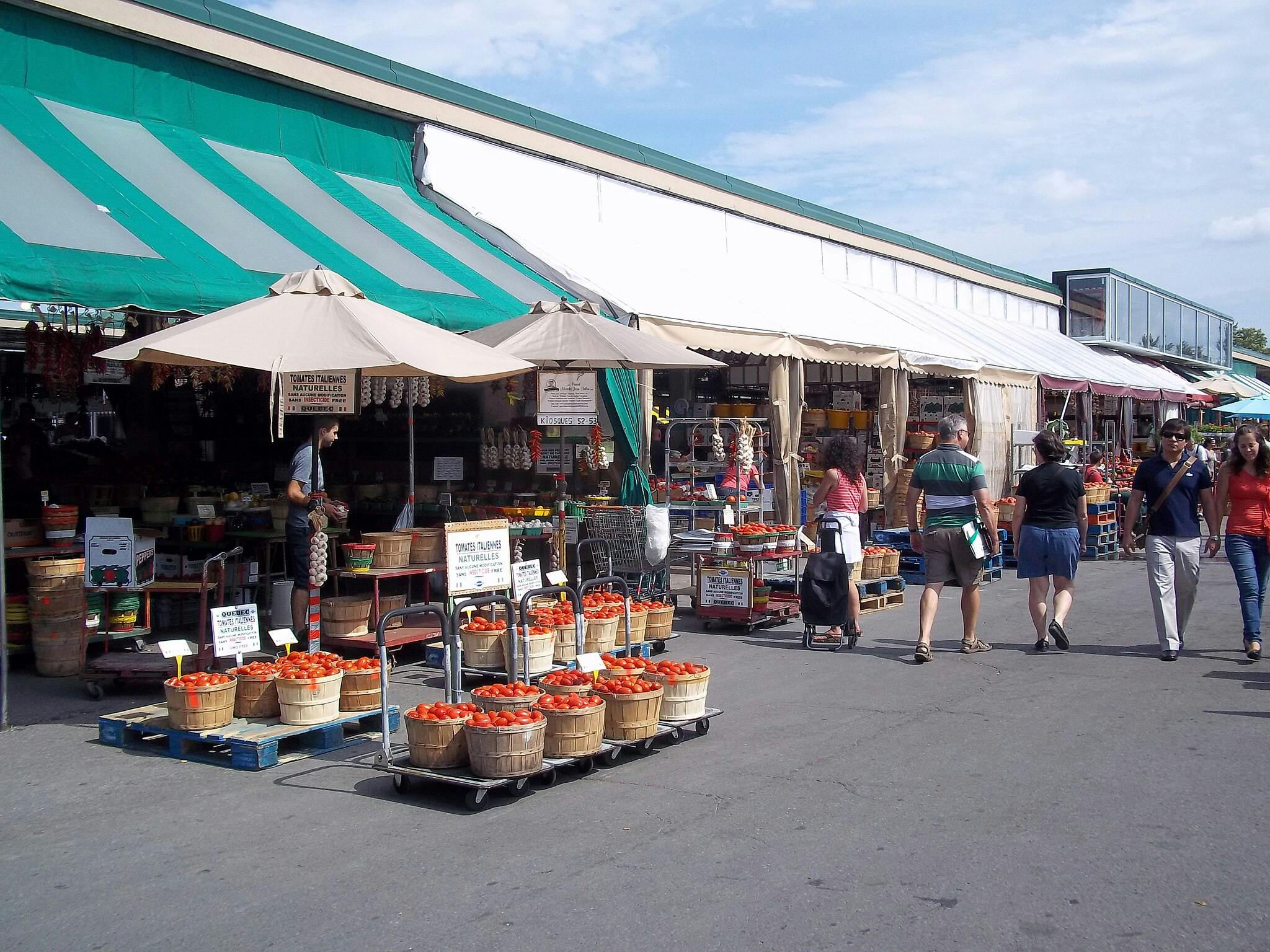 Markt im Freien mit Ständen, die Körbe mit Tomaten unter Regenschirmen verkaufen. Die Menschen gehen an einem sonnigen Tag in Freizeitkleidung vorbei.