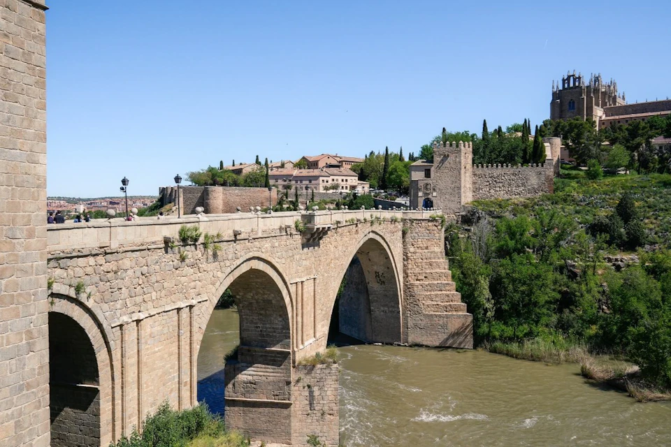 A historic stone bridge crossing a river, surrounded by greenery and buildings under a clear blue sky.