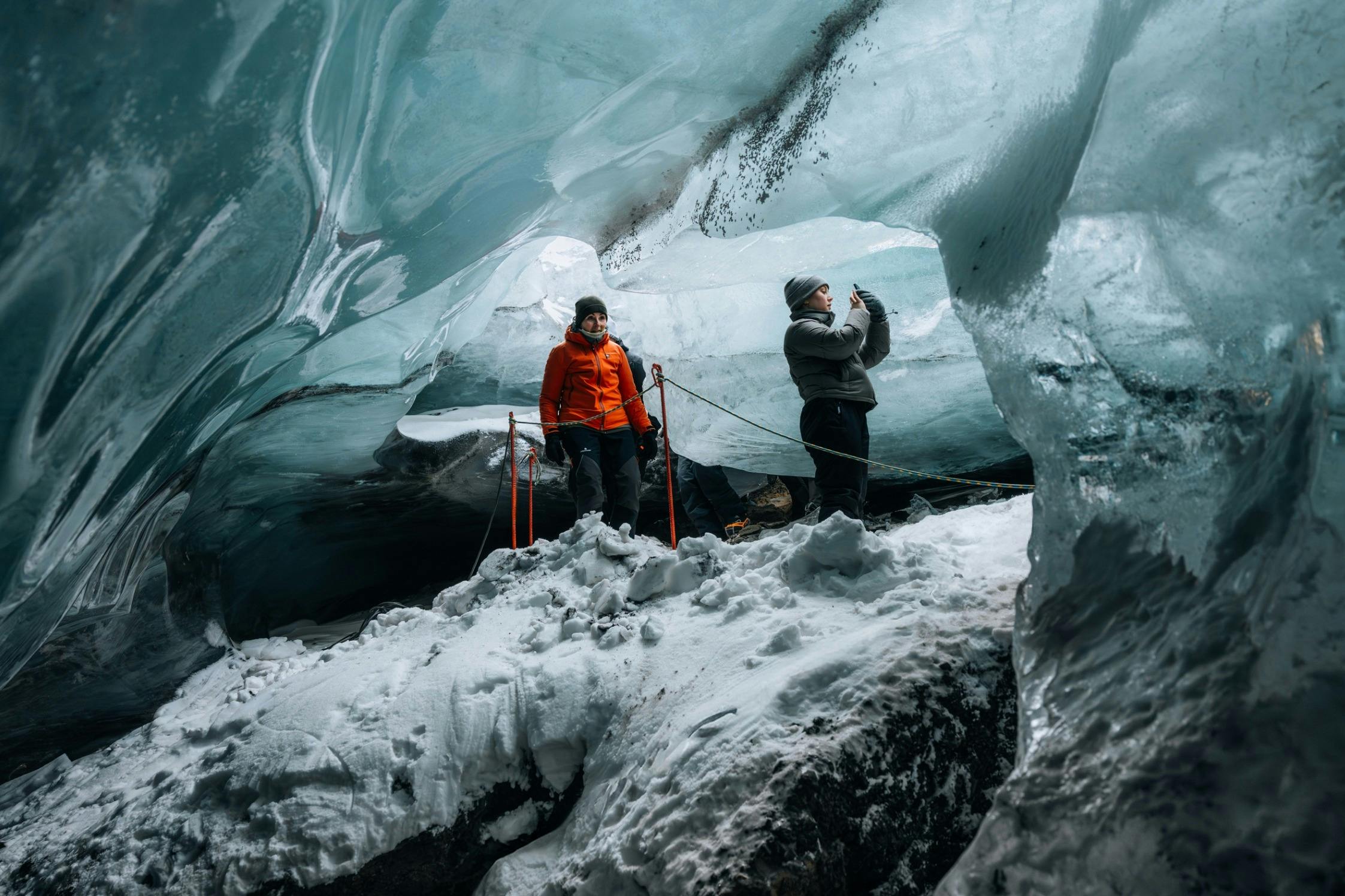 Dva lidé stojící uvnitř ledové jeskyně, jeden v oranžové bundě a druhý v šedé, obklopeni zasněženými a ledovými stěnami.