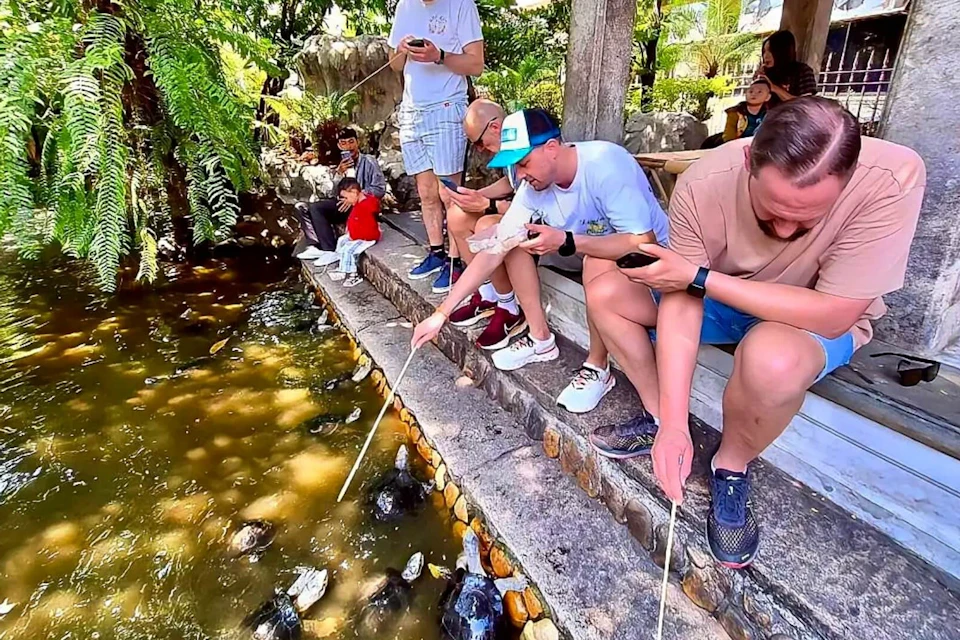 A group of people sitting and standing by a pond feeding fish with sticks under a shaded area in a garden.