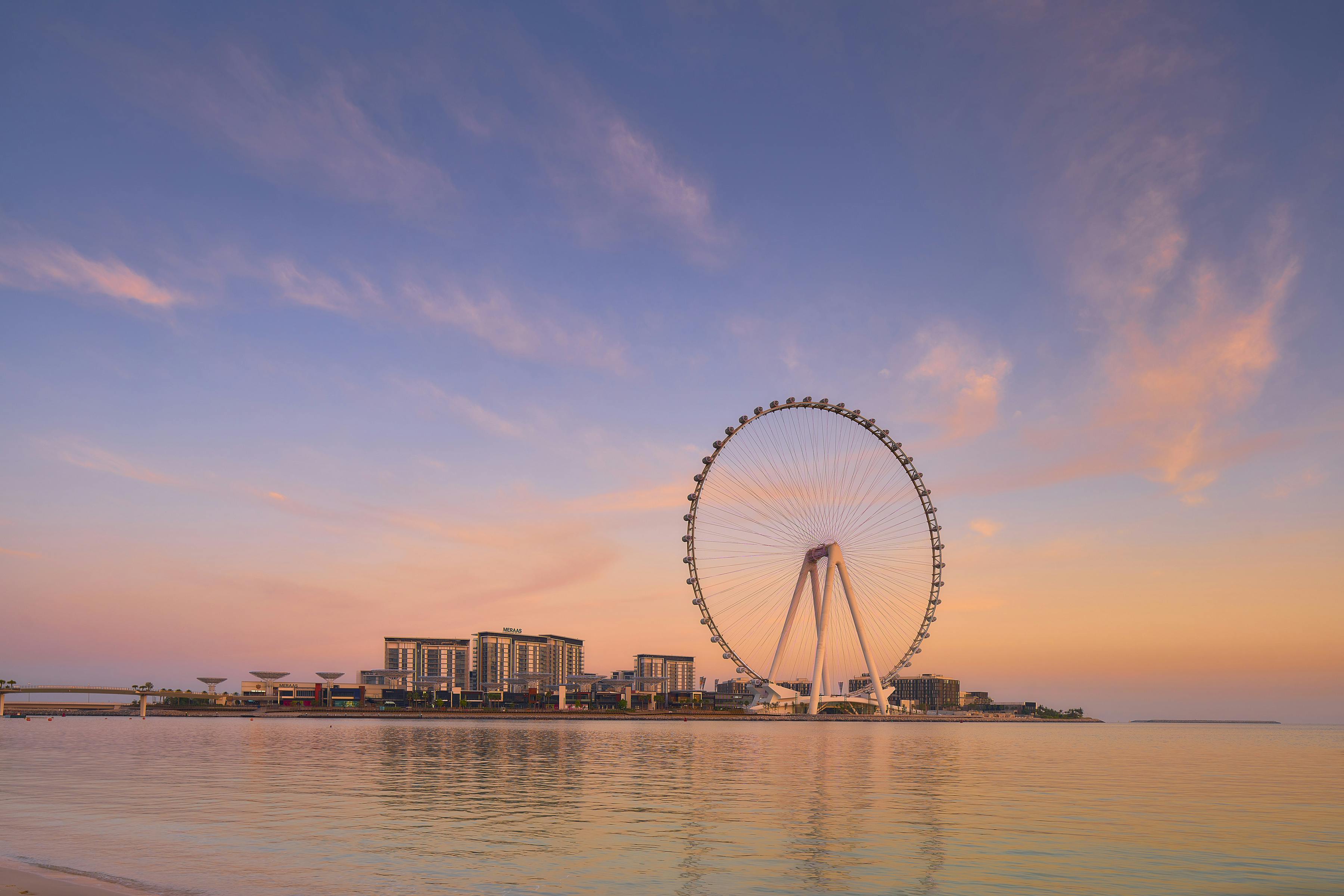 Una grande ruota panoramica ed edifici moderni vicino a un lungomare durante un tramonto colorato con un mare calmo e un cielo pastello.