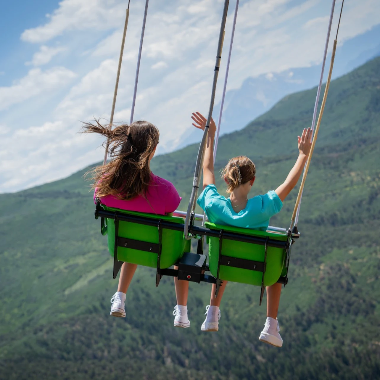 Two people swing high above a mountainous landscape, with one person raising their arms.