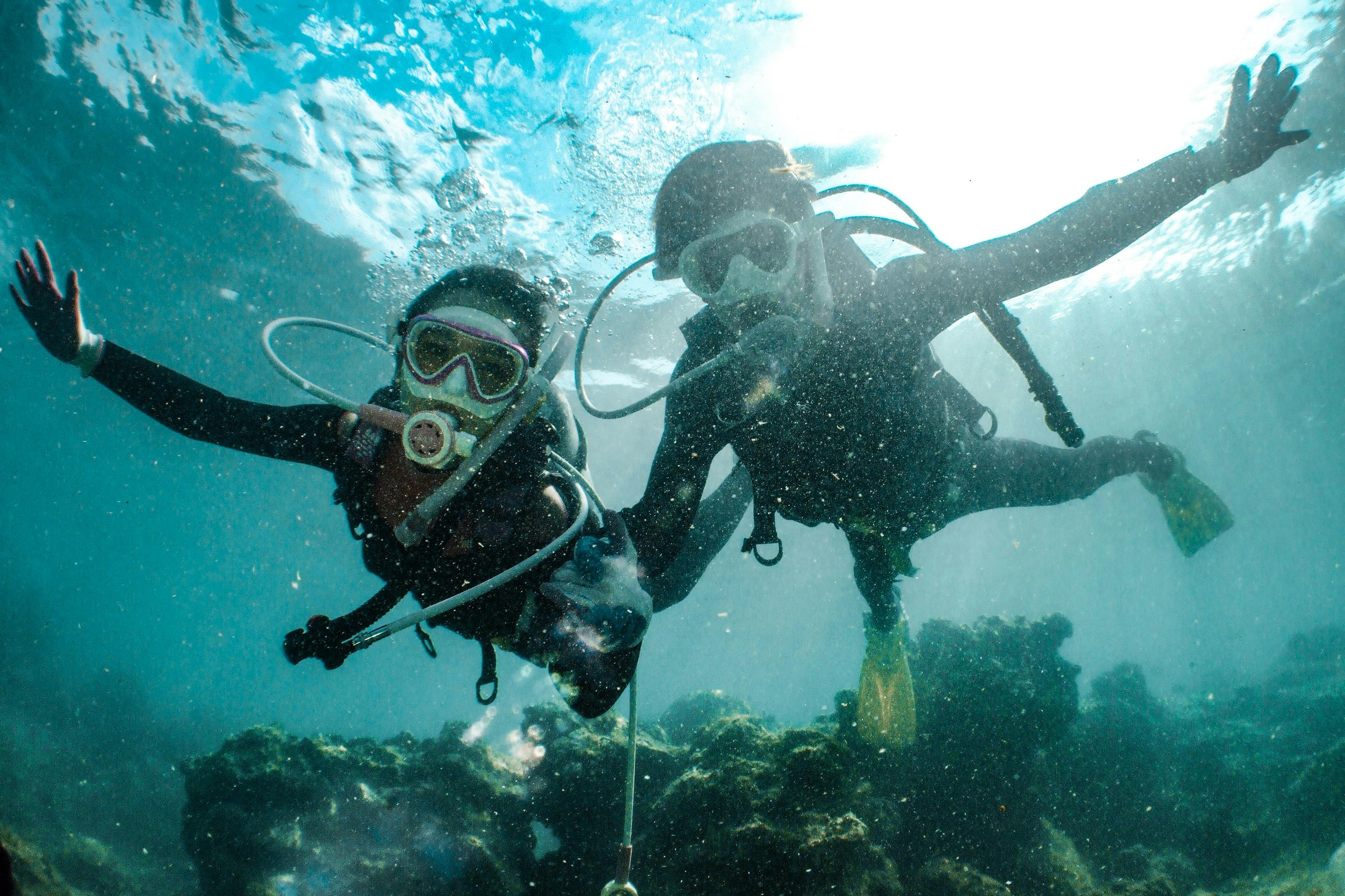 Two scuba divers underwater with gear, masks, and breathing apparatus, swimming above coral reefs with light filtering through the water.