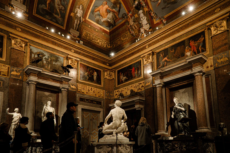 A museum gallery with statues, ornate ceilings, and paintings on the walls. Visitors observe the art and architecture.