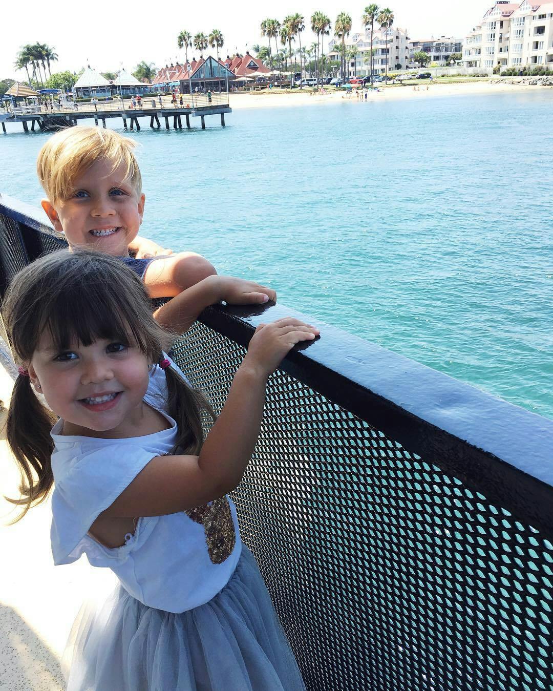 两个微笑的孩子站在栏杆旁，俯瞰平静的蓝色大海，后面有一个码头和建筑物。