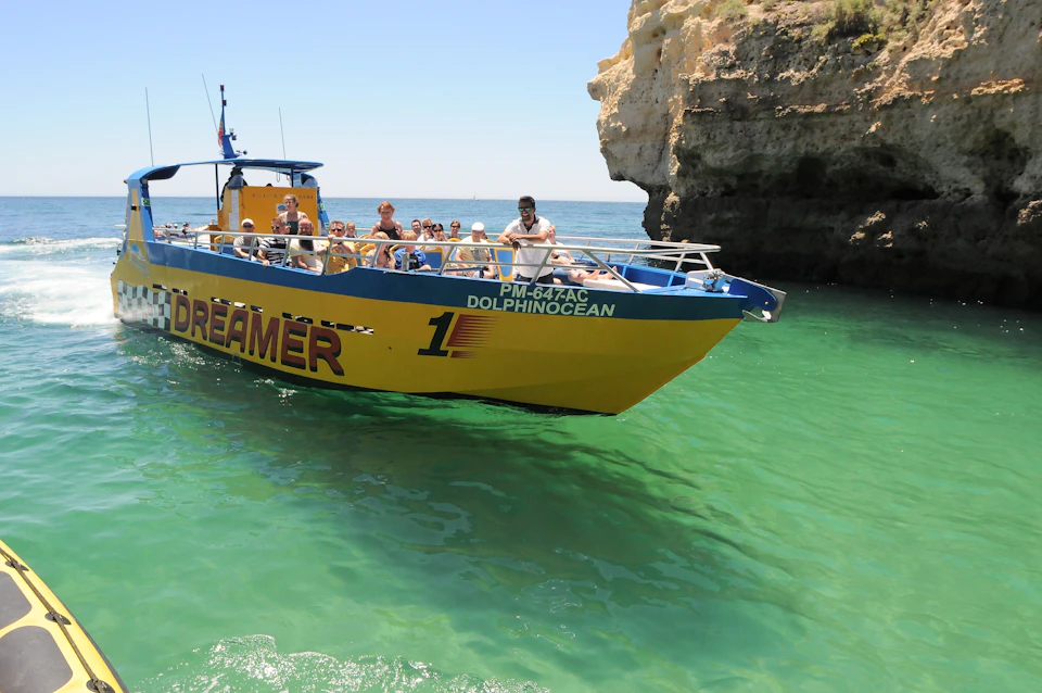 A yellow tour boat named "DREAMER" with passengers sails near a rocky cliff over clear green water on a sunny day.