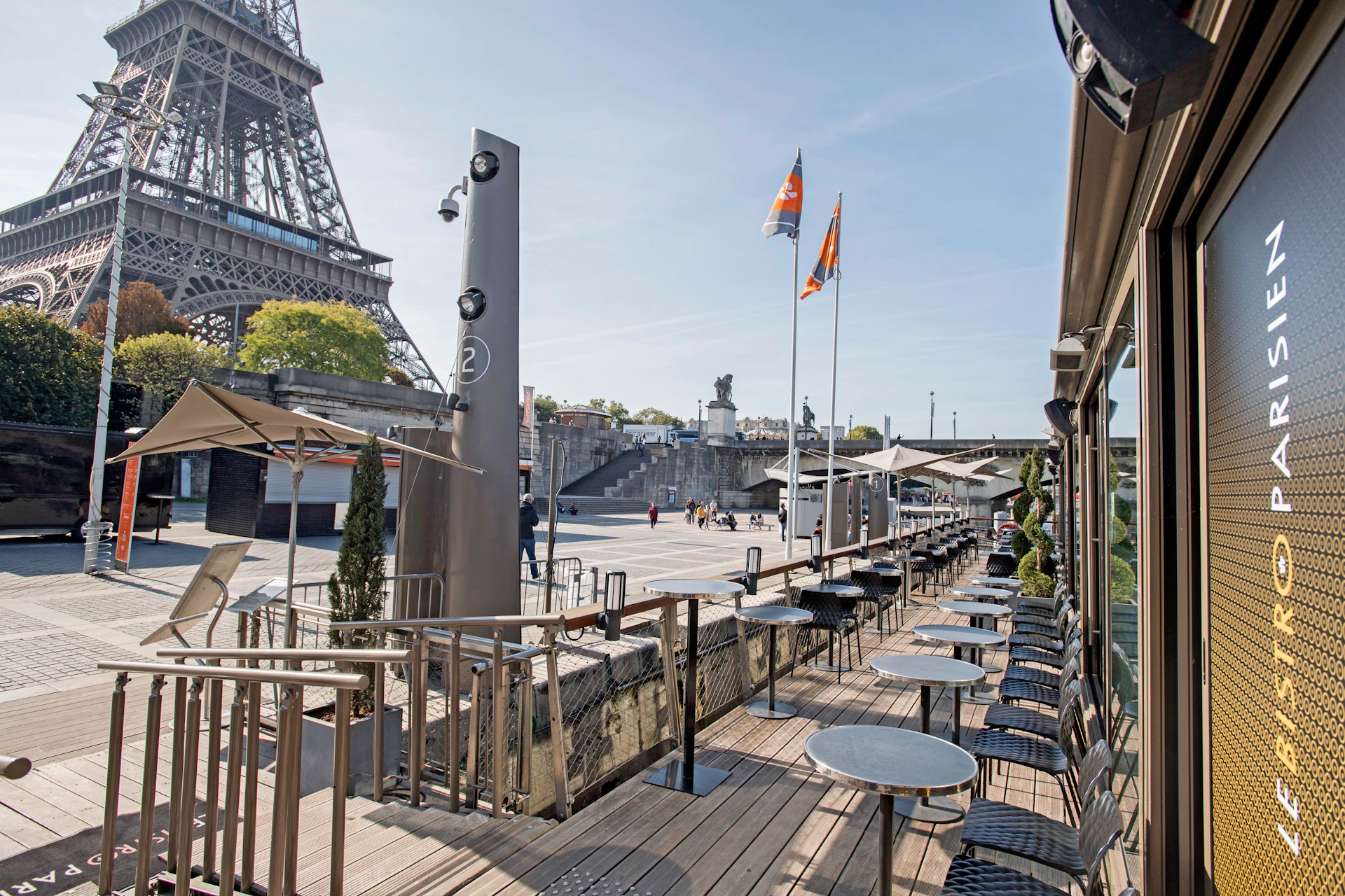Outdoor café seating area with empty tables and chairs beside the Eiffel Tower, flags, and few people in the square.