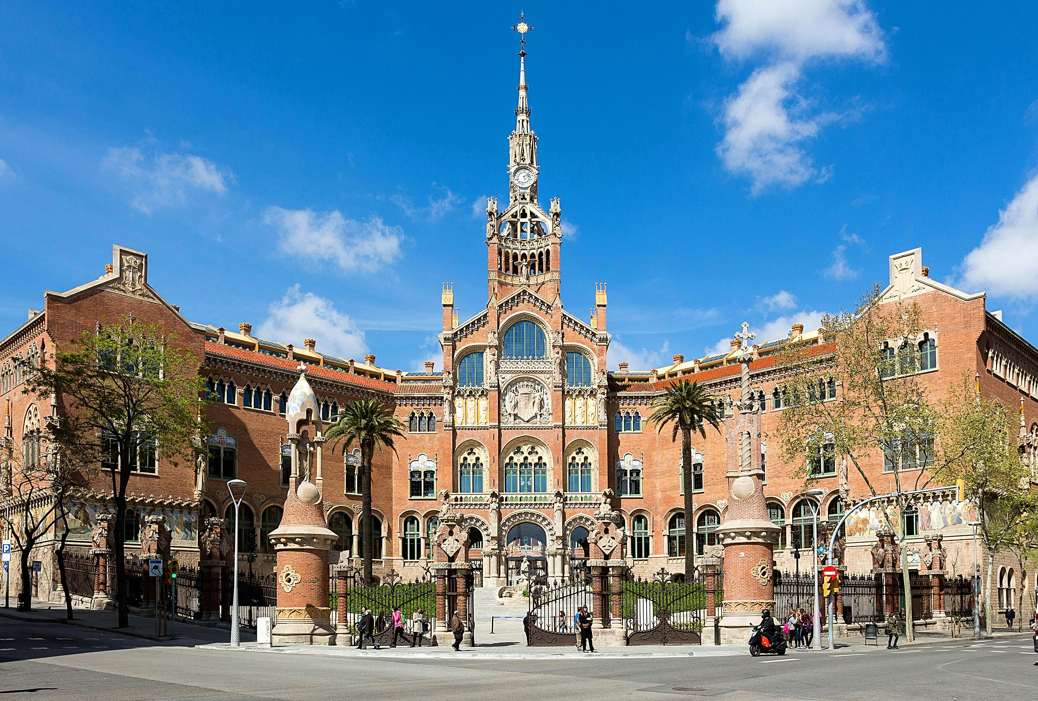 A historic building with intricate architecture and a tall spire, flanked by palm trees, with people walking and a blue sky above.