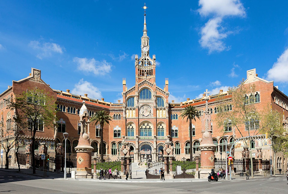 A historic building with intricate architecture and a tall spire, flanked by palm trees, with people walking and a blue sky above.