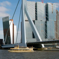 En båt märkt "De Pannenkoekenboot" seglar under en modern bro med höga byggnader i bakgrunden.