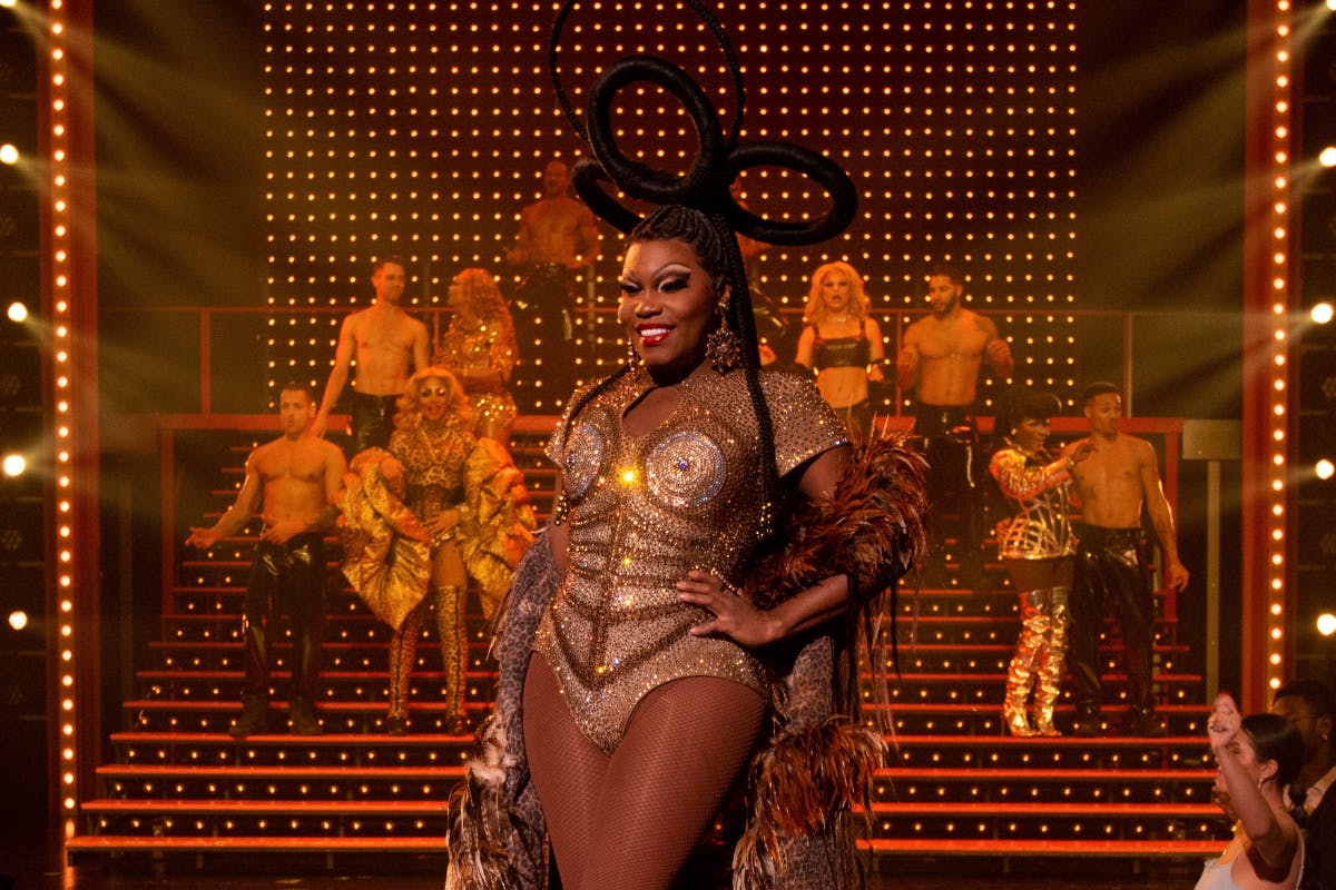 表演者身着金色服装，舞台上有一个大头饰，两侧是五颜六色的服装舞者，背景中有聚光灯。