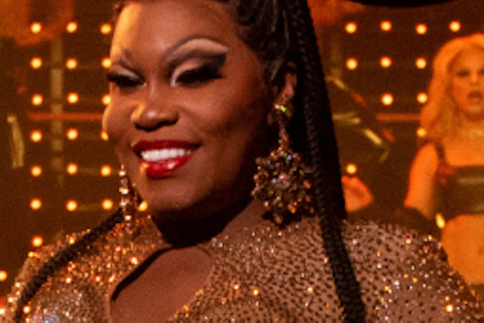 Performer in a gold costume with a large headpiece on stage, flanked by dancers in colorful outfits and spotlights in the background.