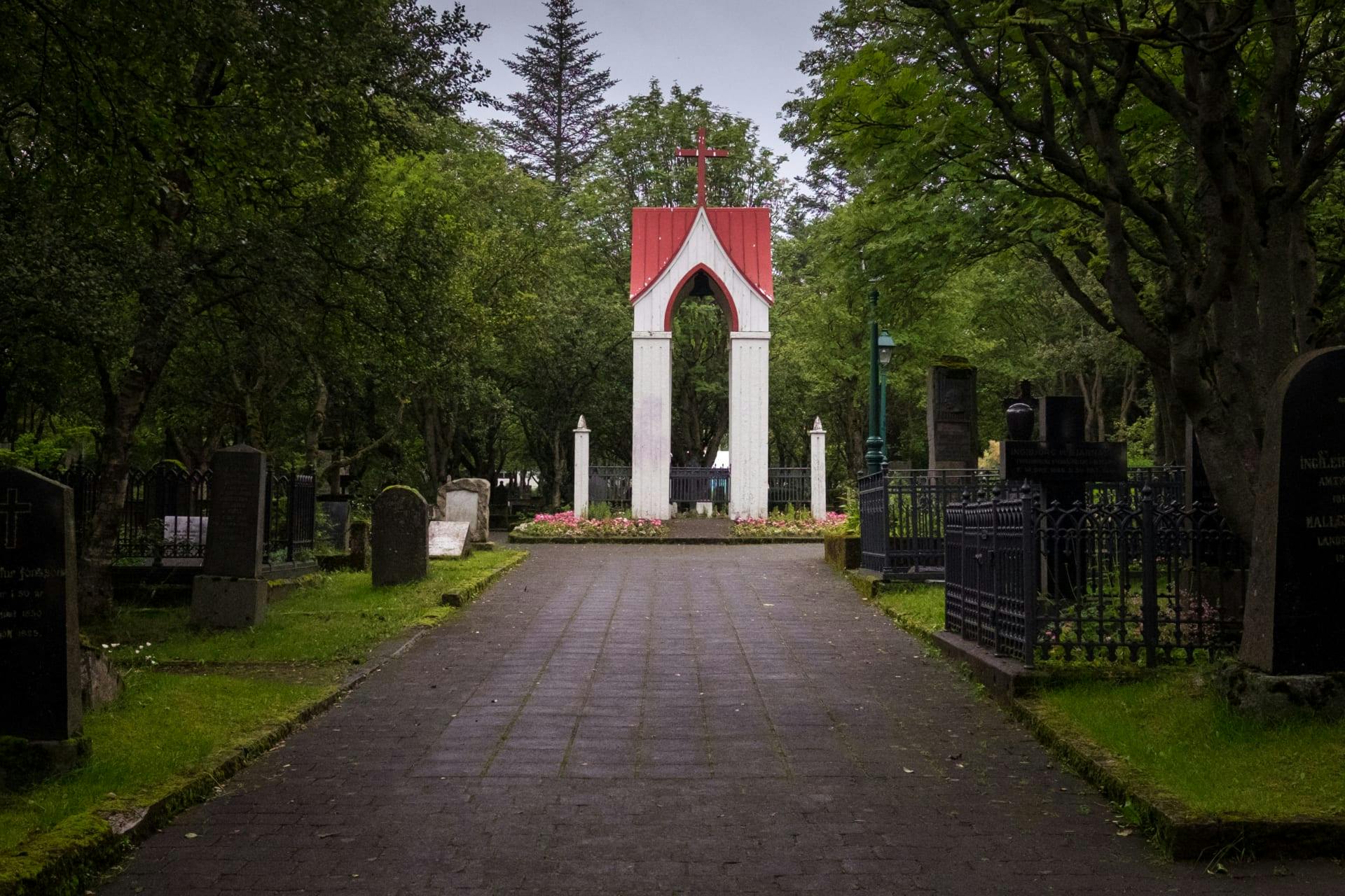 Pathway leading to a white and red archway with a cross, surrounded by tombstones and trees in a cemetery.