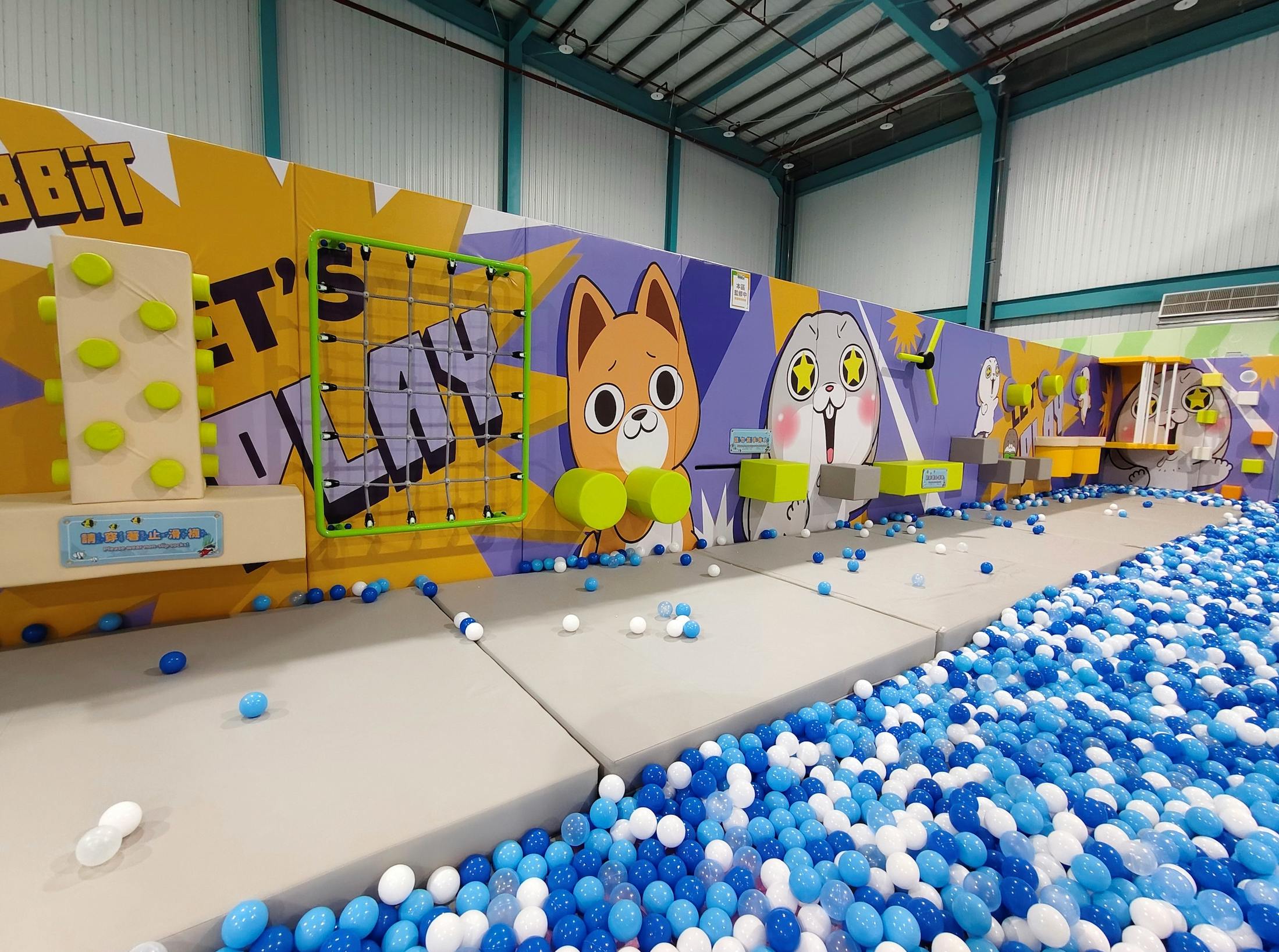 Indoor play area with colorful cartoon-themed wall art, a ball pit with blue and white balls, and padded flooring.