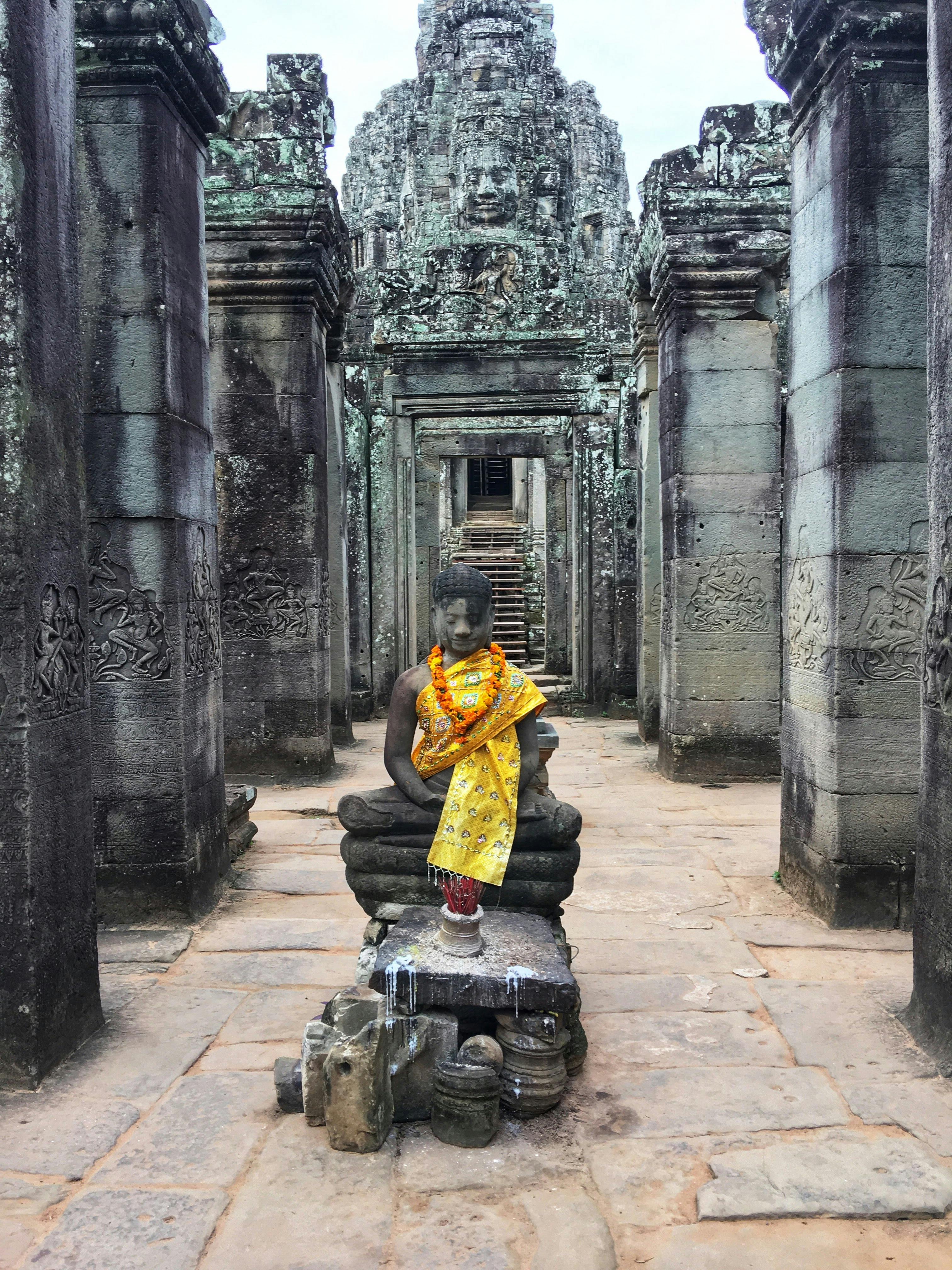 Ancient stone temple hallway with a seated Buddha statue adorned with yellow cloth and flower garlands in the foreground.
