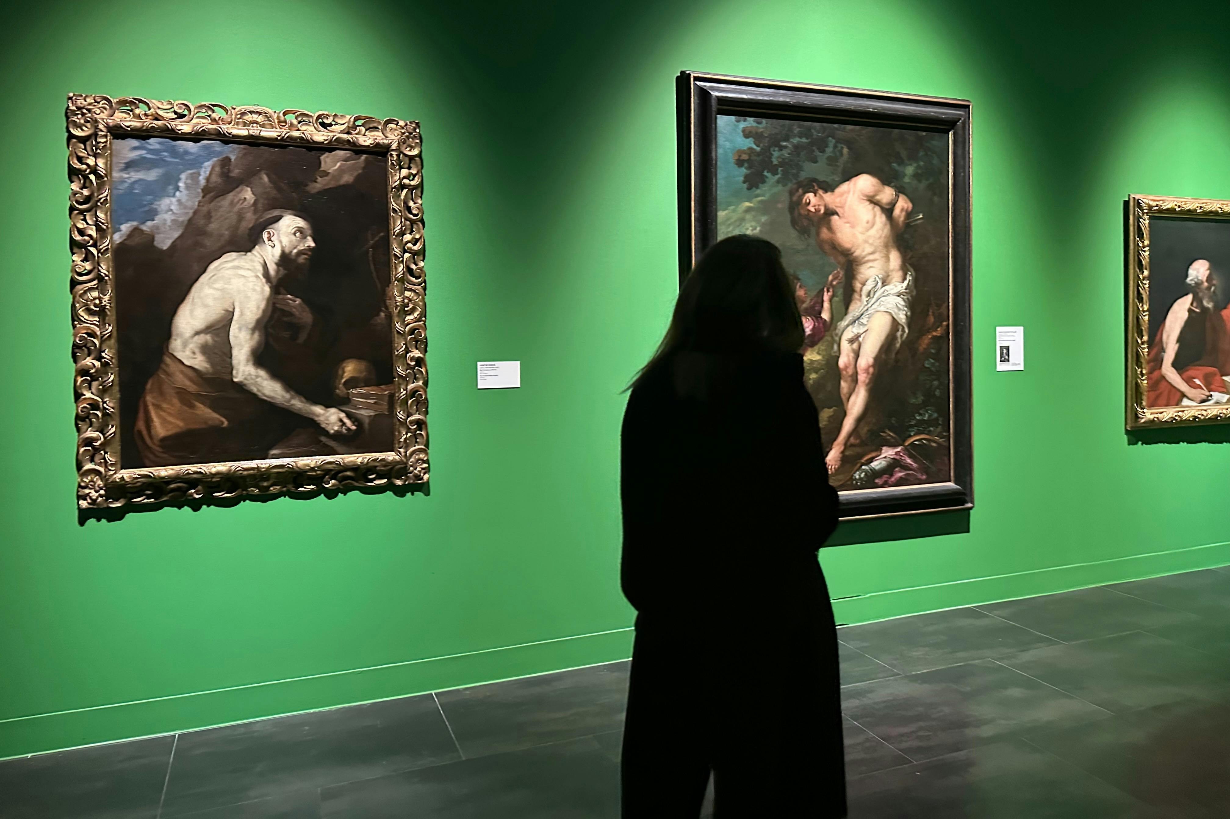 Uma pessoa em silhueta contra paredes verdes de galerias com quadros emoldurados; um retrata uma figura amarrada a uma árvore, outro um homem sentado.