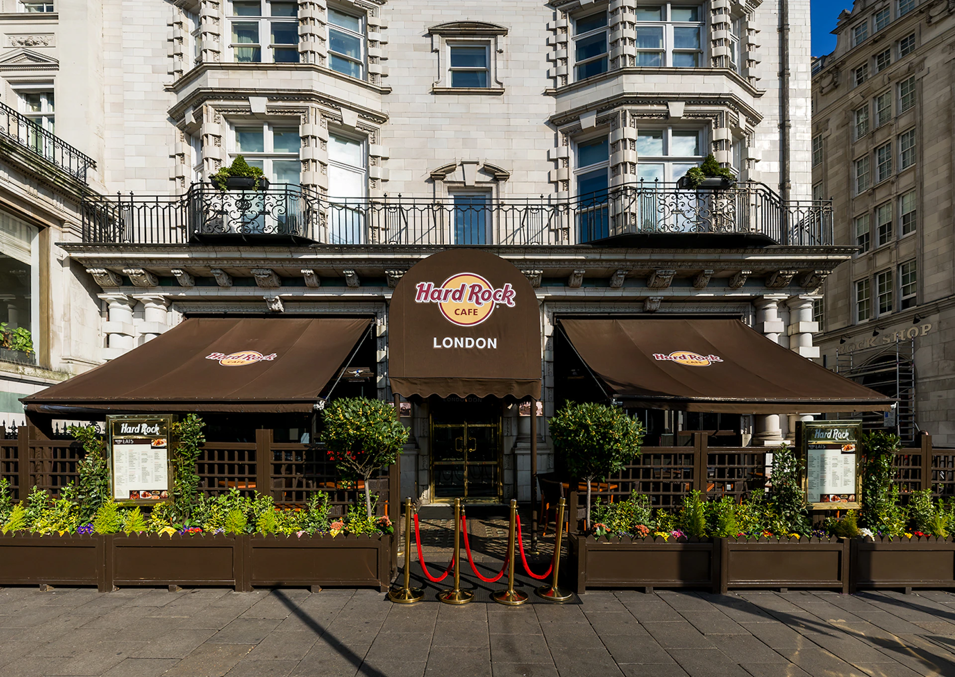 Exterior of Hard Rock Cafe London with brown awnings, potted plants, and red ropes leading to the entrance.