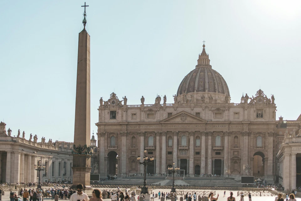 A view of a large basilica with a dome and an obelisk in the foreground, surrounded by people in an open plaza.
