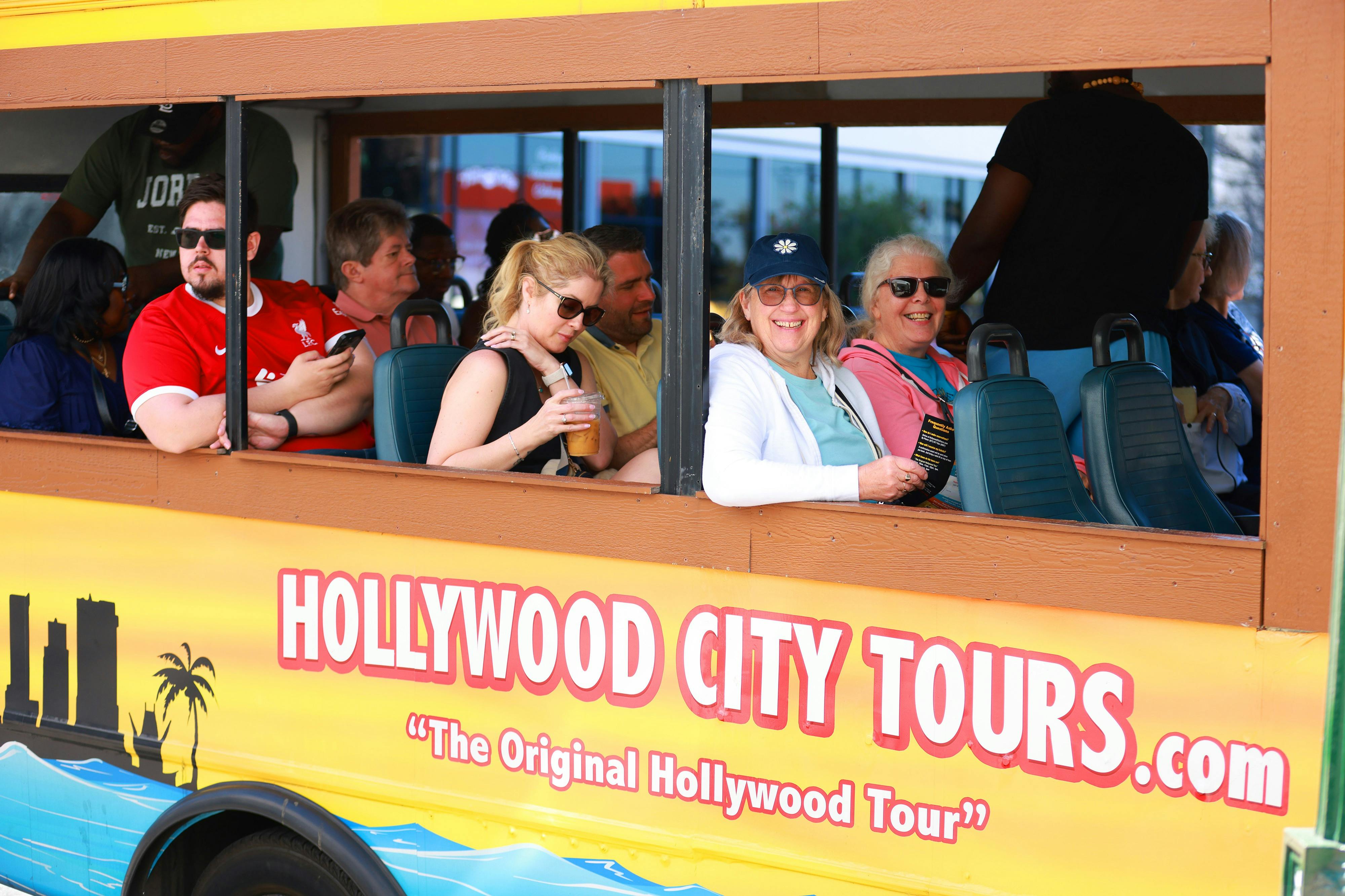 Turistes somrients s'asseuen en un bus per fer "Hollywood City Tours". L'autobús té les finestres obertes i el nom del recorregut es mostra al costat.