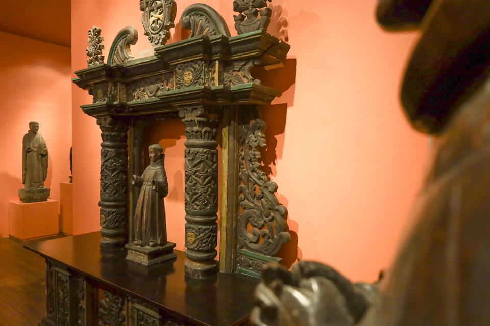 Ornate wooden sculpture with intricate carvings, featuring columns and a human figurine, displayed against a pink wall.