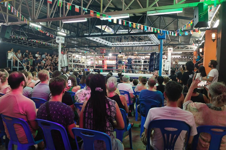 A crowd of spectators seated in front of a boxing ring, with two fighters inside under a lit canopy in an indoor venue.