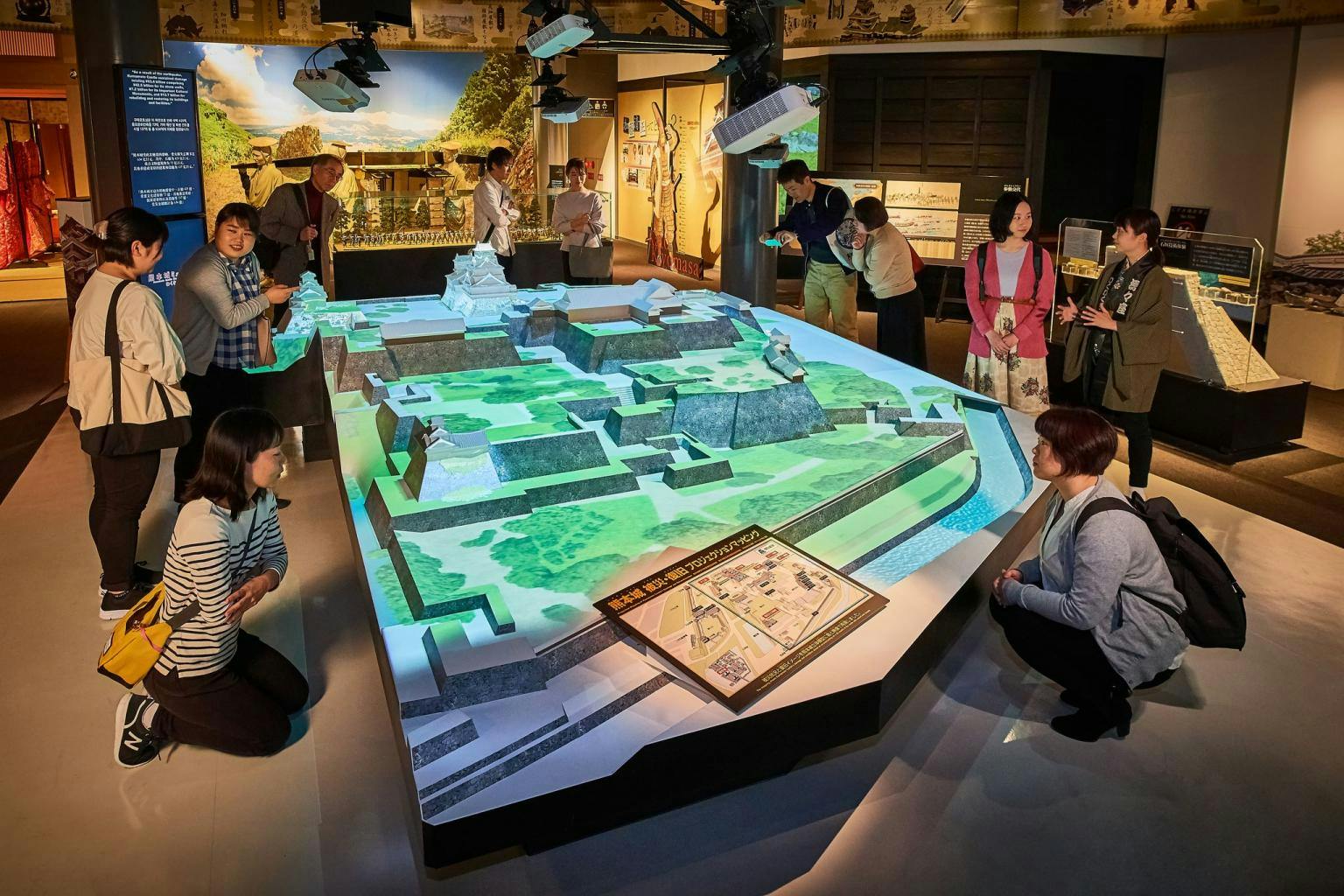 People observing a detailed 3D model of a historical castle in a museum exhibit under focused lighting.