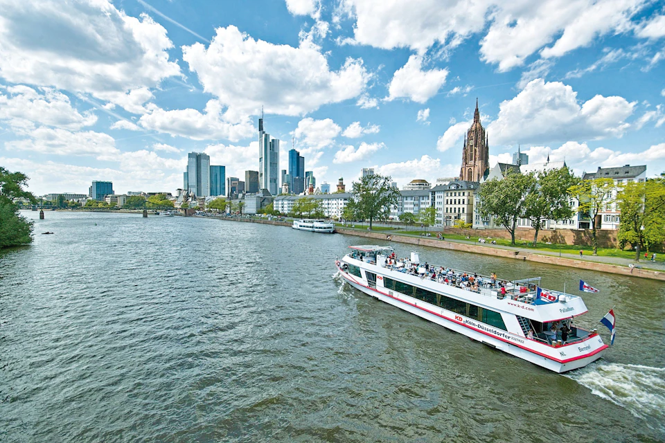 A river scene with a boat carrying passengers, a city skyline with modern and historic buildings, and a blue sky with clouds.