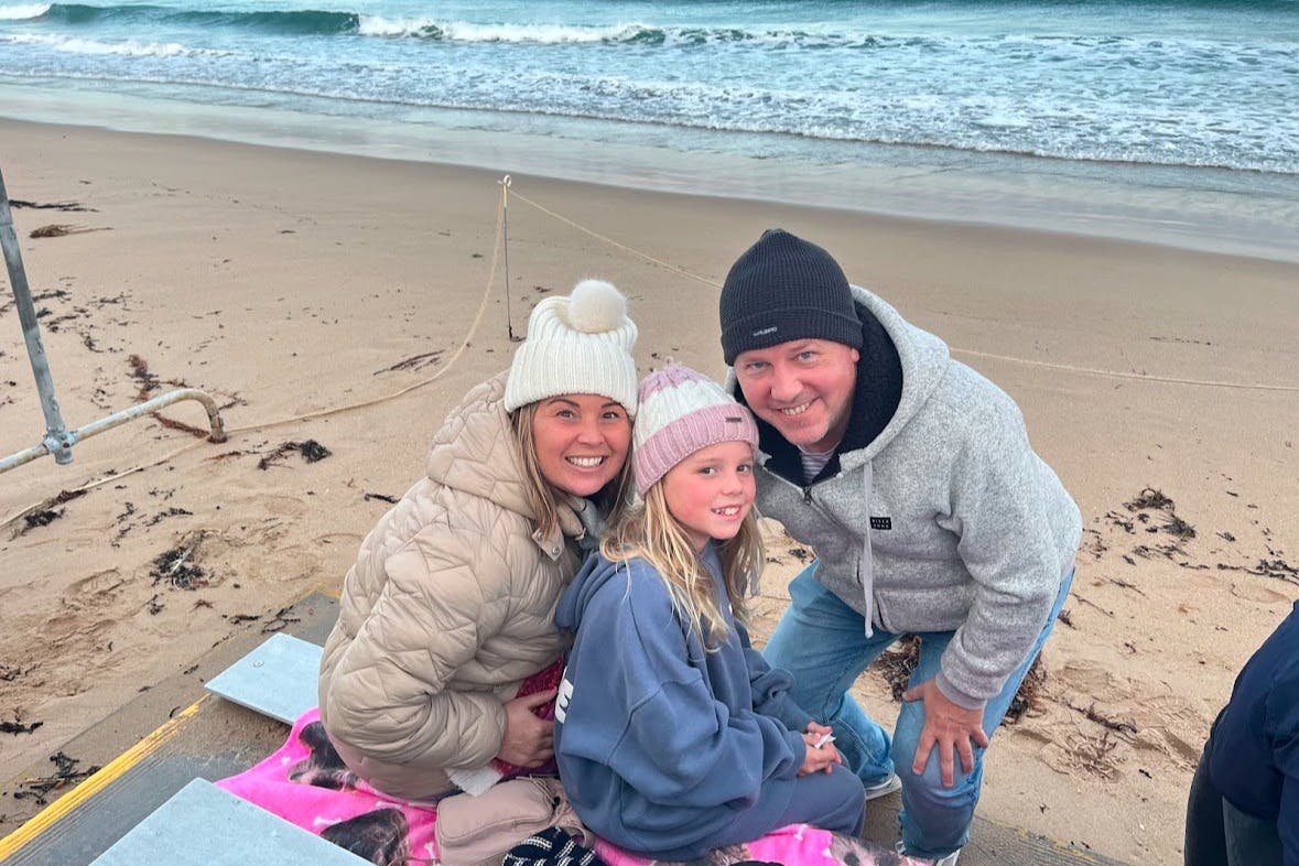 A family of three, bundled in winter clothing and hats, smiles while sitting on a beach blanket near the shore.