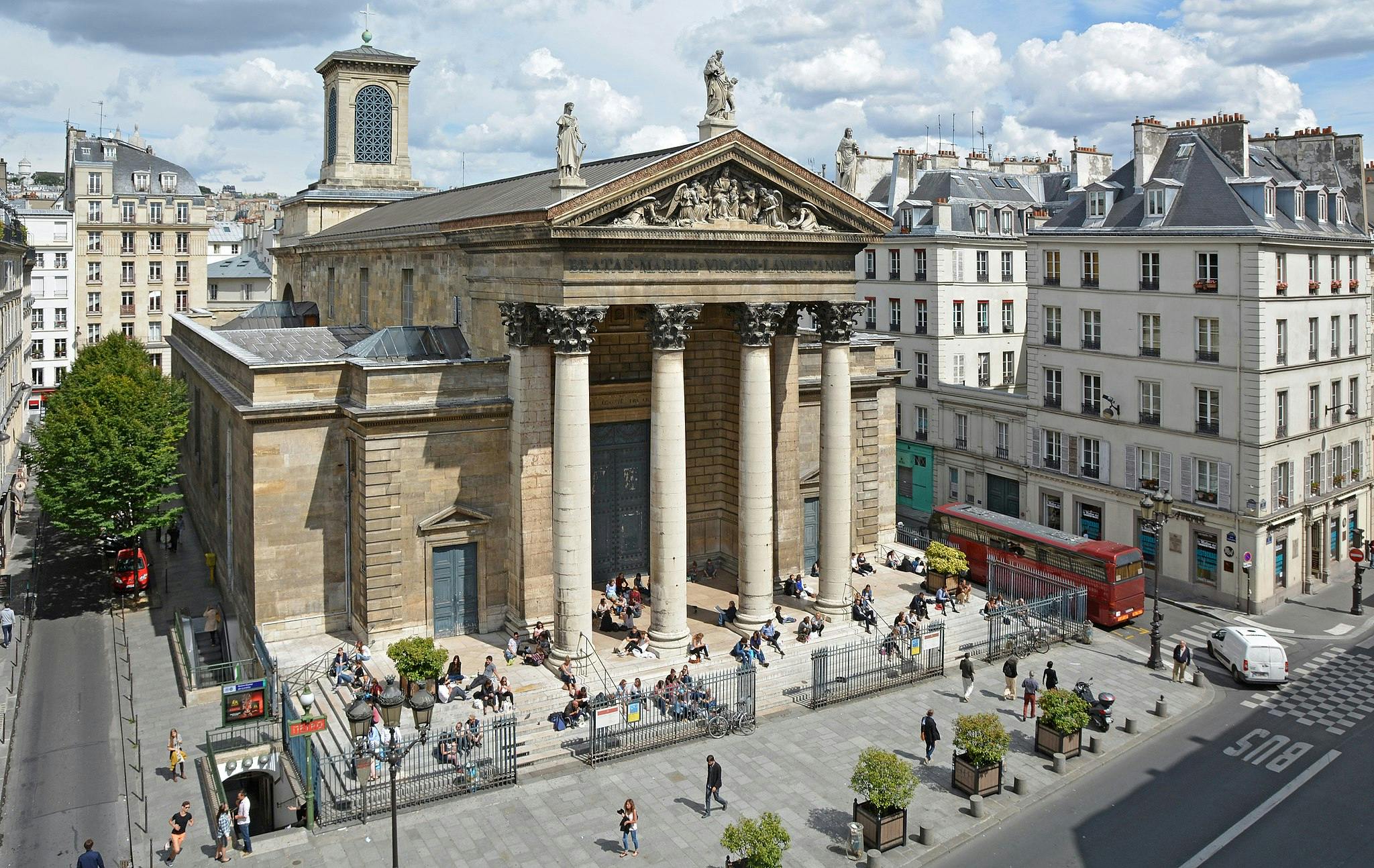 People sitting and walking near a historic stone building with tall columns; surrounding it are urban streets and buildings.