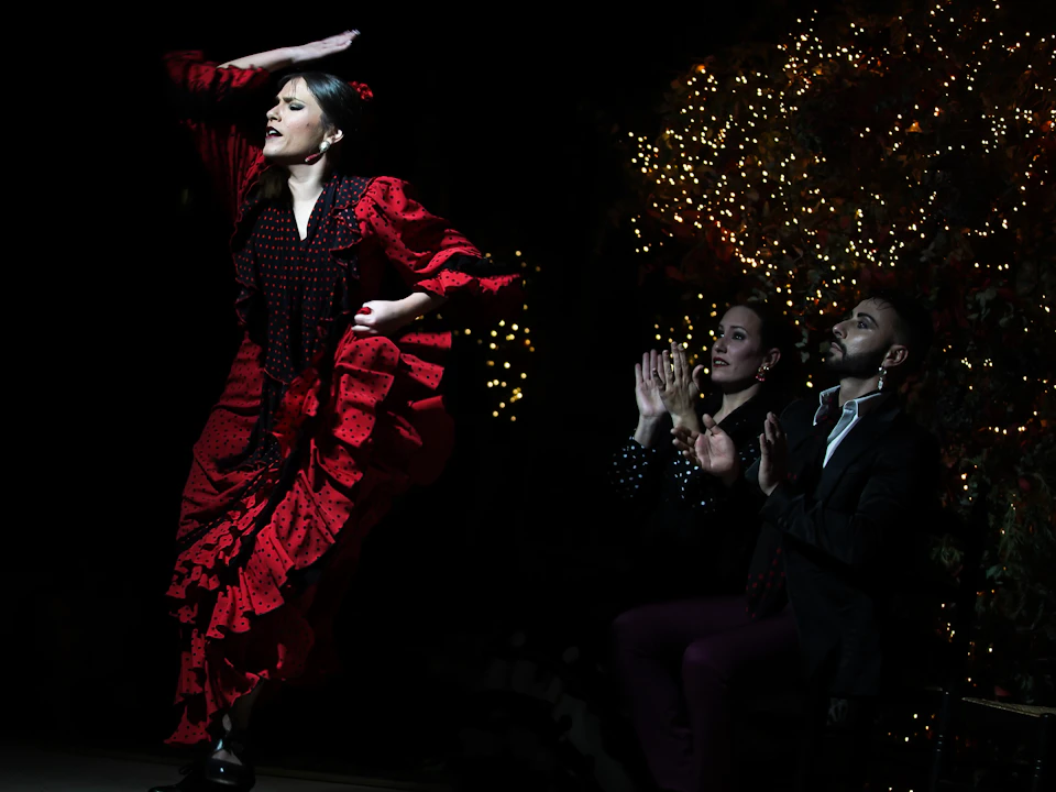 Flamenco dancer in a red dress performs passionately on stage, while two seated people in dark attire clap in rhythm. Twinkling lights decorate the background.