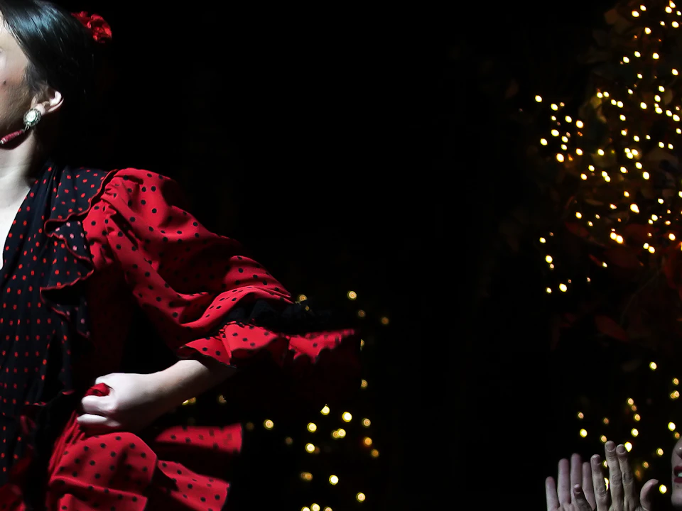 Flamenco dancer in a red dress performs passionately on stage, while two seated people in dark attire clap in rhythm. Twinkling lights decorate the background.
