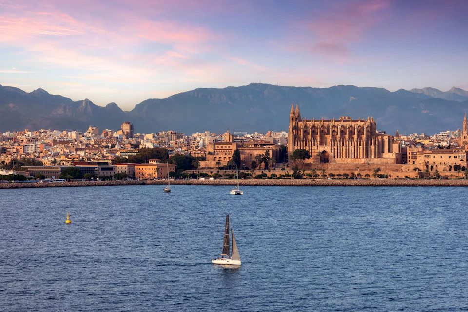 A coastal cityscape with a cathedral and mountains in the background, a sailboat in the foreground on a calm sea at sunset.
