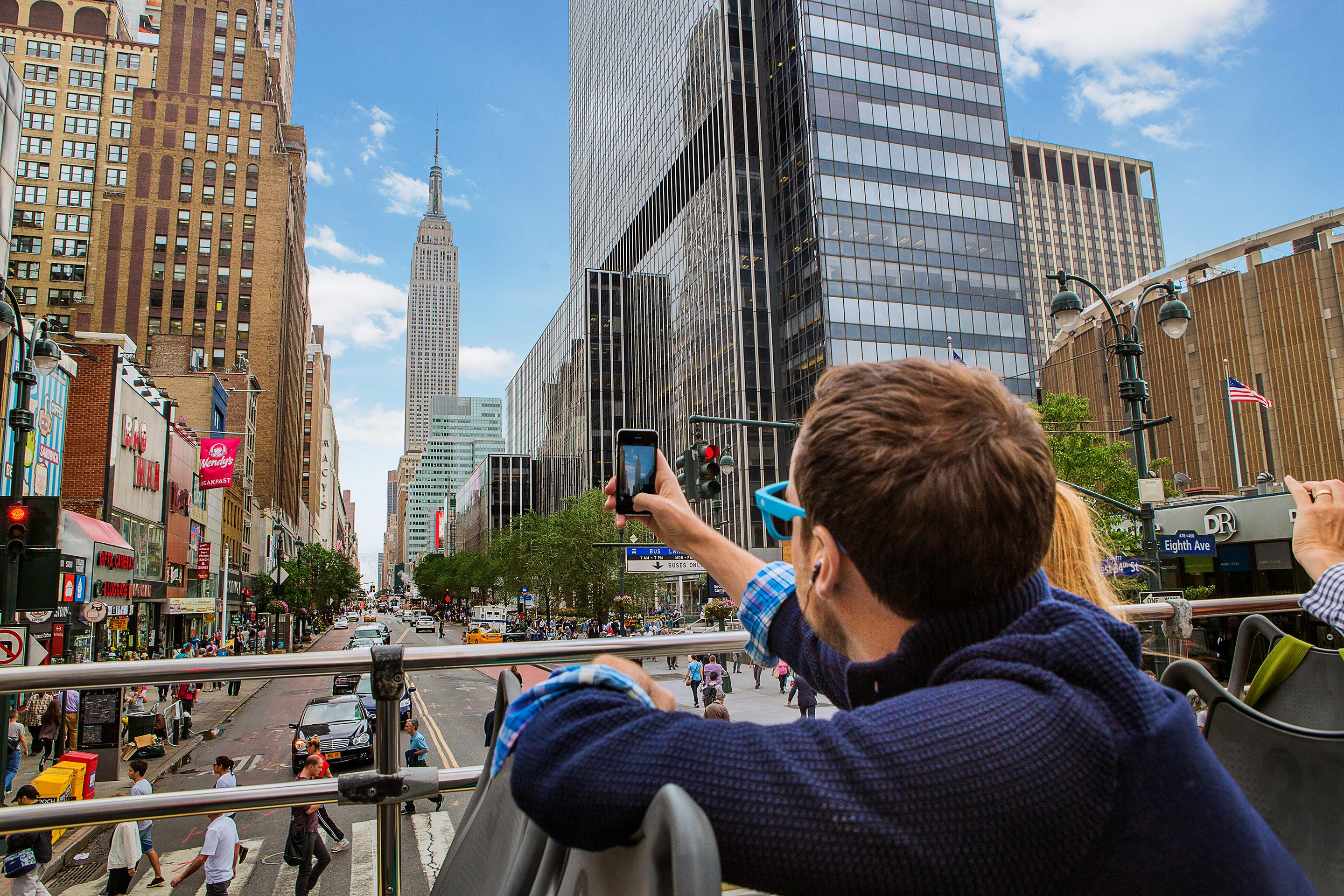 A person takes a photo of the Empire State Building from an open-top bus in a busy New York City street.