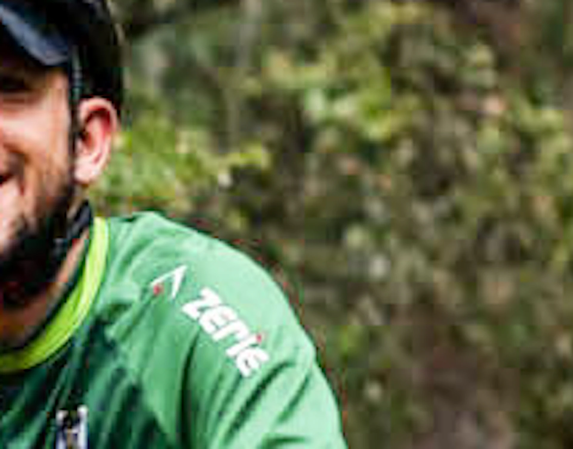Three people riding bicycles on a dirt path through a forest, smiling and wearing helmets. The background is lush with trees and greenery.