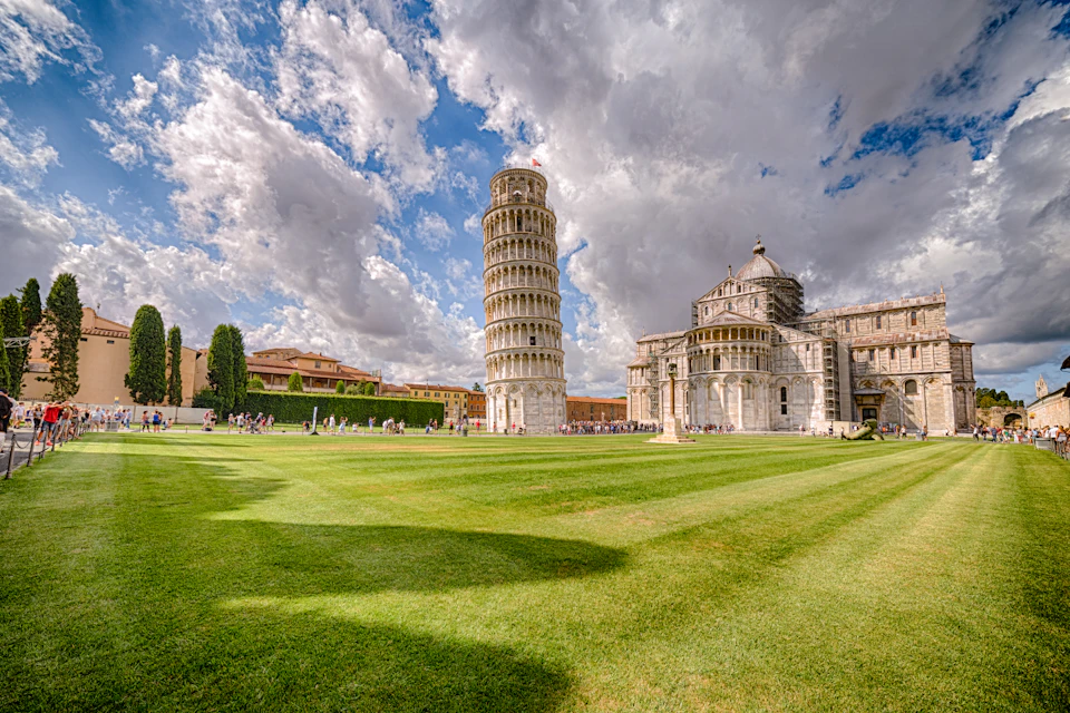 Leaning Tower of Pisa and adjacent cathedral under a cloudy sky, with a grassy foreground and scattered tourists.