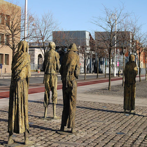 Fem vittrade bronsstatyer av människor som står på en kullerstensbelagd trottoar, omgiven av lövlösa träd, urbana byggnader i bakgrunden.