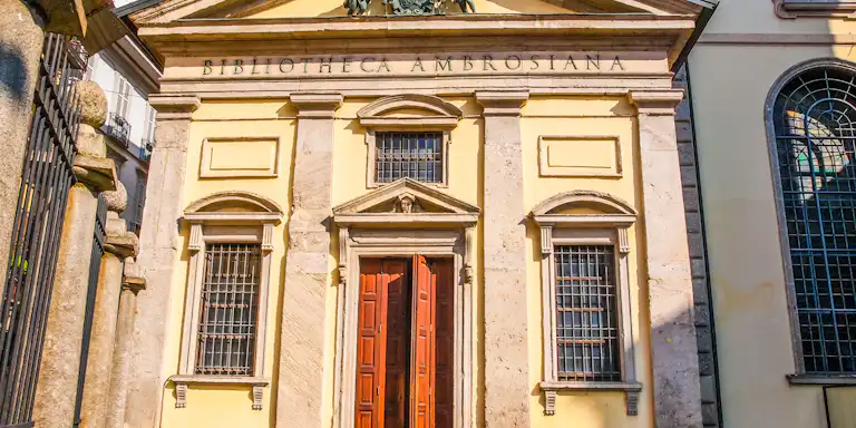 鮮やかな青空の下、木製の扉の上に「Bibliotheca Ambrosiana」と刻まれた華やかな建物。