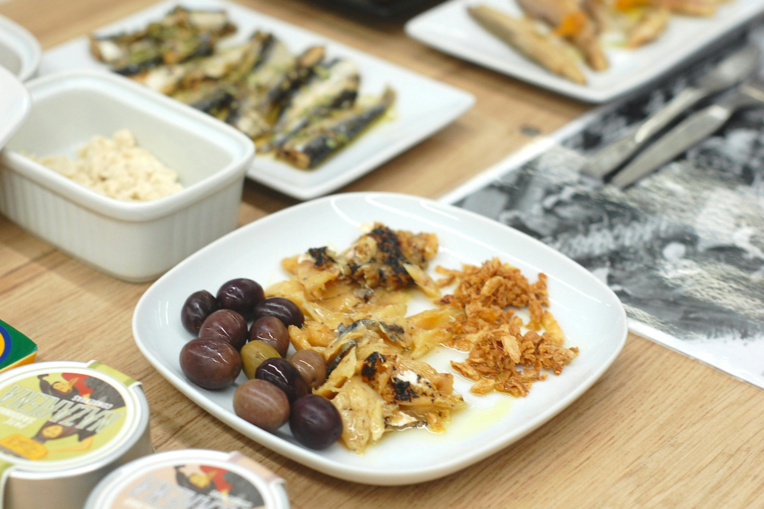 Bord met zwarte olijven, gegrilde stukjes vis en knapperige uitjes op een houten tafel. Andere gerechten zijn gedeeltelijk zichtbaar op de achtergrond.