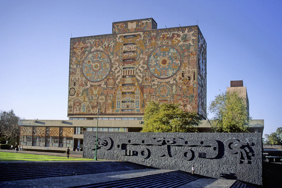 A large, colorful mural decorates the façade of a tall building, with a stone sculpture in the foreground and trees to the side.