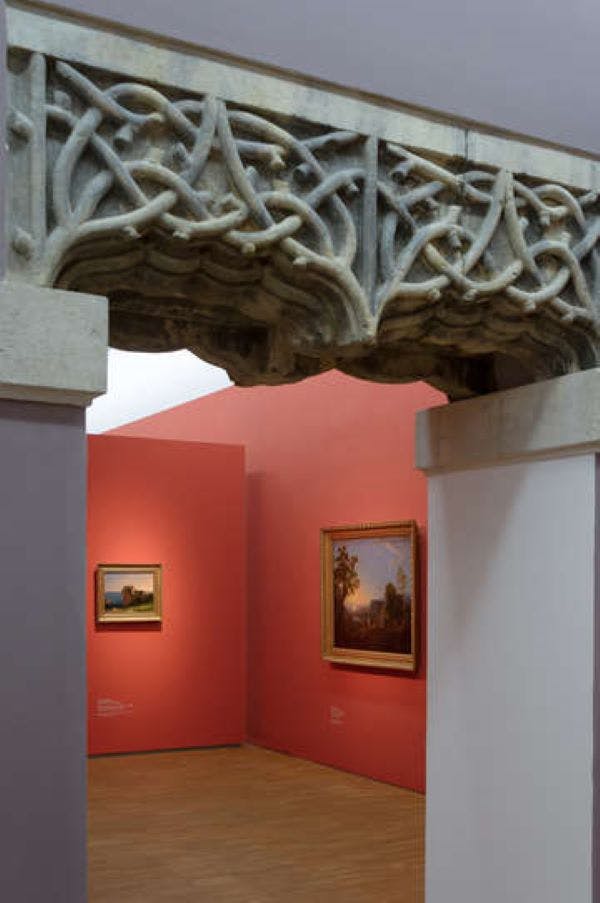 Decorative stone archway with intricate carvings framing a gallery scene with two paintings on coral-colored walls.