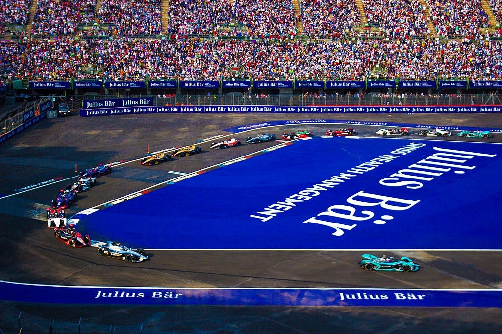 Auto da corsa che affrontano una curva su una pista con il brand "Julius Bär". Le tribune sono riempite da una grande folla di spettatori.