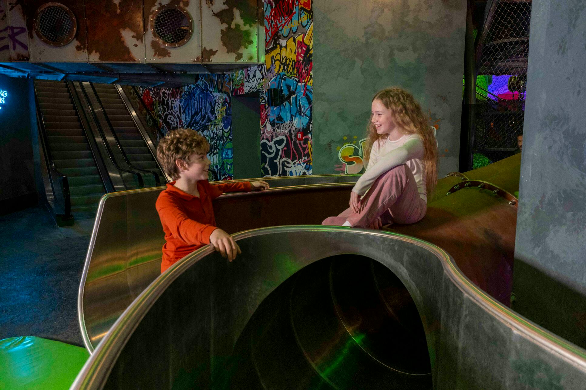 Twee kinderen in interactie in een overdekte speelruimte met glijbanen en kleurrijke met graffiti bedekte muren.