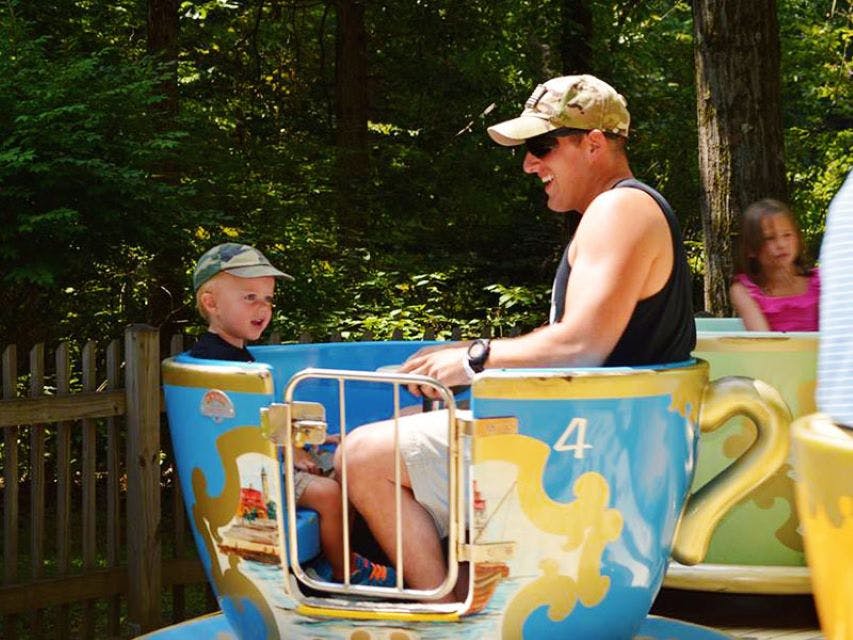 Een man en een jongen rijden in een blauw theekopje in een pretpark, met een meisje in een roze shirt op de achtergrond.