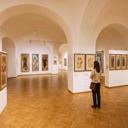 En person ser inramade konstverk som visas på väggarna i ett galleri med välvda tak och trägolv.